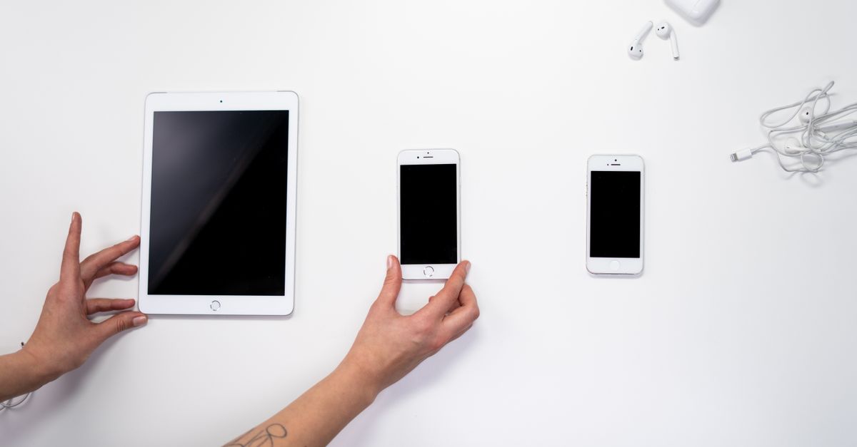 Top view of hands arranging an iPad, iPhones, and earphones on a white surface. Perfect for tech-related imagery.